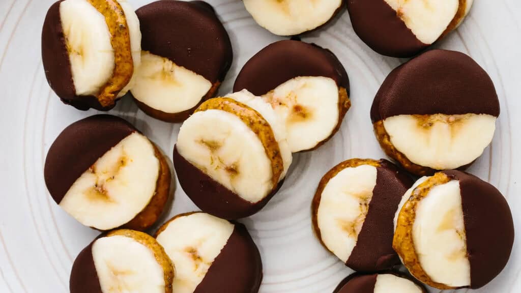 A plate of bite-sized snacks, each consisting of a banana slice sandwiched with a layer of nut butter and partially dipped in dark chocolate. Perfect for those exploring new snack recipes, these treats are arranged in a random pattern on a white surface.