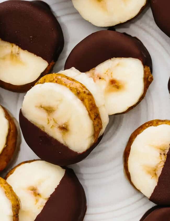 A plate of bite-sized snacks, each consisting of a banana slice sandwiched with a layer of nut butter and partially dipped in dark chocolate. Perfect for those exploring new snack recipes, these treats are arranged in a random pattern on a white surface.