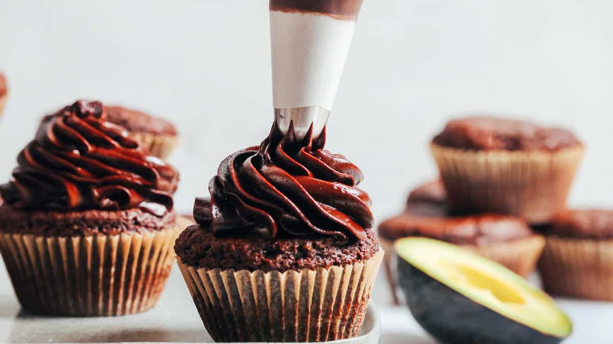 A close-up of a chocolate cupcake being decorated with swirls of chocolate frosting from a piping bag. More frosted cupcakes and a halved avocado, known for its health benefits, are blurred in the background. The scene is set on a light-colored surface, perfect for capturing delicious recipes.