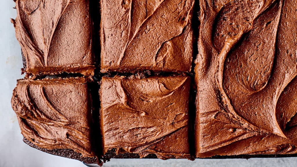 Close-up of a chocolate cake topped with rich, creamy chocolate frosting, cut into neat squares. The swirled texture of the frosting is prominently visible, giving a sense of the cake's moist and indulgent nature—perfect for those looking for decadent sheet cake recipes.