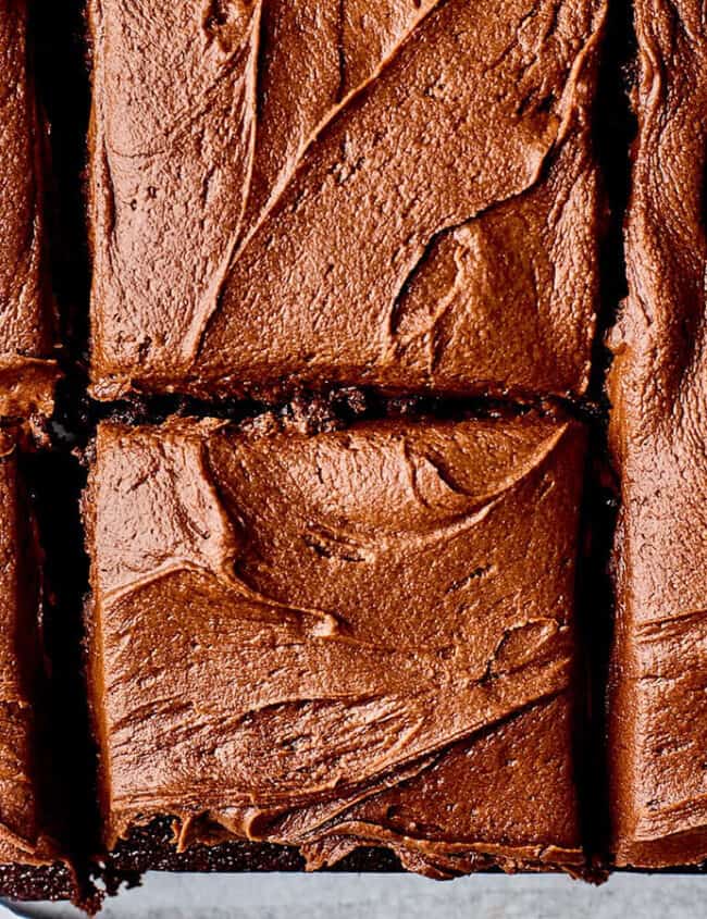 Close-up of a chocolate cake topped with rich, creamy chocolate frosting, cut into neat squares. The swirled texture of the frosting is prominently visible, giving a sense of the cake's moist and indulgent nature—perfect for those looking for decadent sheet cake recipes.