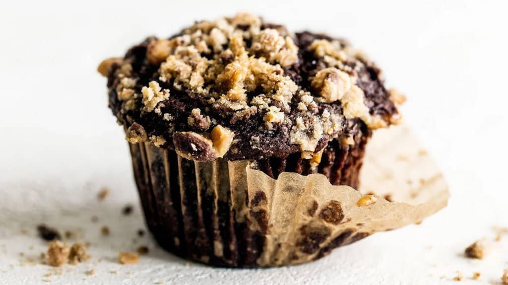 A close-up of a chocolate muffin in a brown paper liner. The muffin has a crumbly topping with chunks of nuts and sugar, and some crumbs are scattered on the white surface below. The liner is partially unwrapped, revealing the moist texture of the muffin—a perfect addition to your chocolate recipes collection.