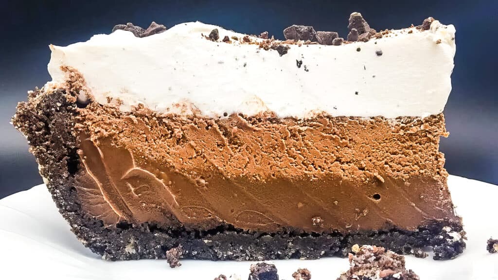 A close-up of a slice of chocolate pie with a crumbly dark chocolate crust, smooth chocolate mousse layer, and a thick, fluffy layer of whipped cream on top. This delectable dessert could easily become your favorite among chocolate recipes. The pie is set against a dark background, highlighting the contrast of textures and colors.