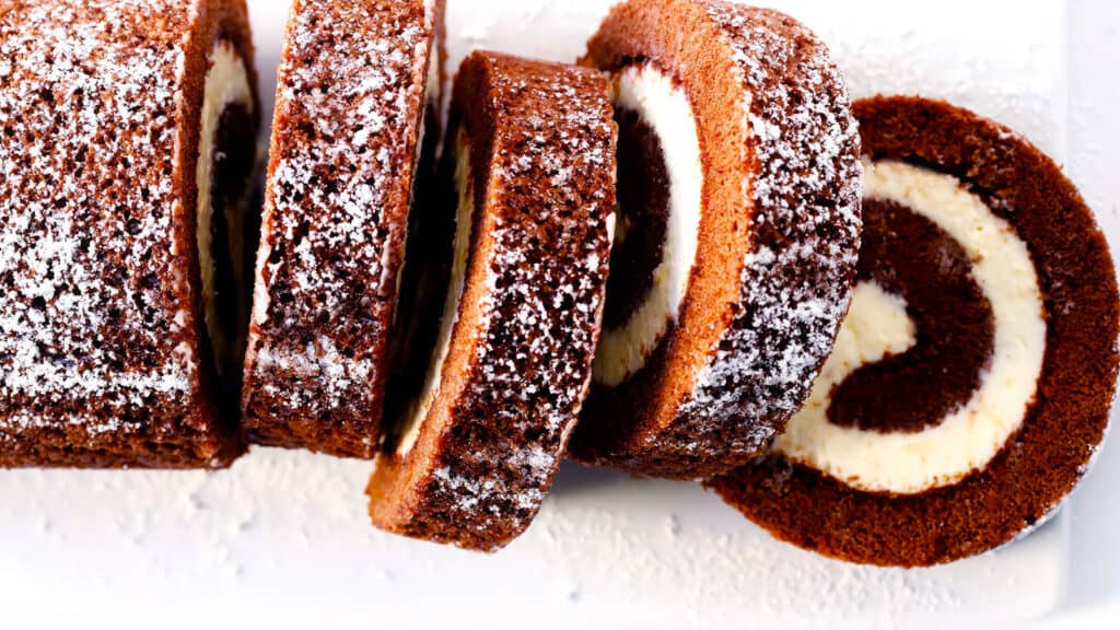 A chocolate Swiss roll cake, a classic in Chocolate Recipes, is dusted with powdered sugar and sliced into multiple pieces, revealing the spiral pattern of the creamy filling inside. The cake has a rich, dark chocolate appearance, with the white filling providing a beautiful contrast.