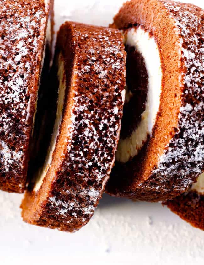 A chocolate Swiss roll cake, a classic in Chocolate Recipes, is dusted with powdered sugar and sliced into multiple pieces, revealing the spiral pattern of the creamy filling inside. The cake has a rich, dark chocolate appearance, with the white filling providing a beautiful contrast.