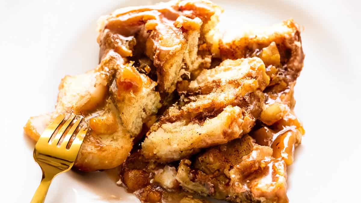 A close-up of a dessert dish featuring pieces of bread pudding drizzled with caramel sauce. The pudding is topped with chunks of cooked apples and a sprinkle of spices. A gold fork is partially inserted into the pudding, ready to be savored—a perfect addition to your collection of pancake recipes.