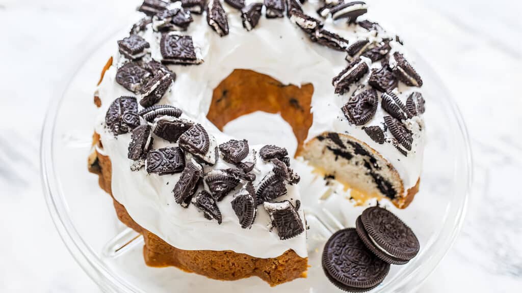 A bundt cake topped with white frosting and crushed chocolate sandwich cookies is displayed on a clear glass cake stand. A slice has been cut from the cake, revealing more cookie pieces inside. Two chocolate sandwich cookies sit next to the cake on the stand, perfect for those exploring Bundt Cake Recipes.