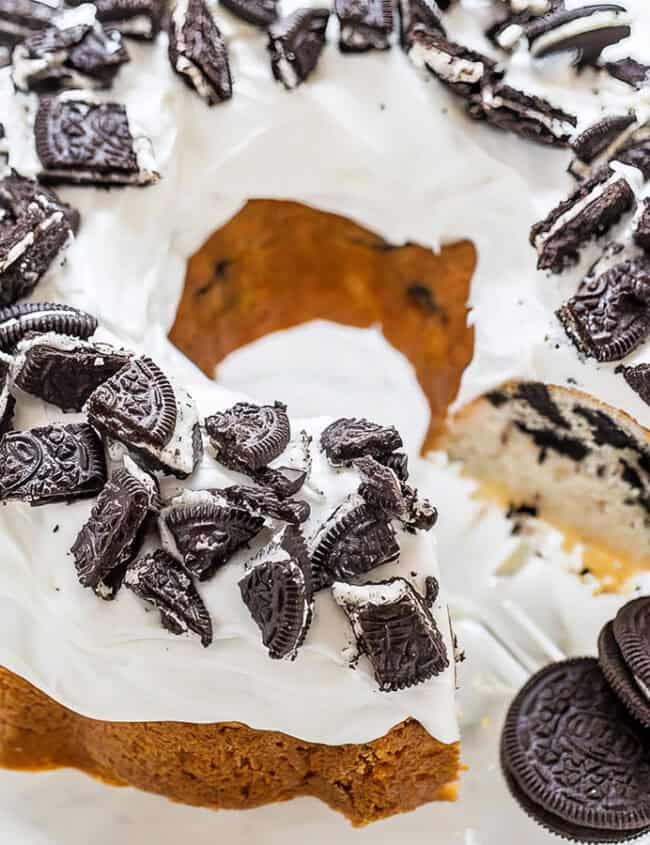 A bundt cake topped with white frosting and crushed chocolate sandwich cookies is displayed on a clear glass cake stand. A slice has been cut from the cake, revealing more cookie pieces inside. Two chocolate sandwich cookies sit next to the cake on the stand, perfect for those exploring Bundt Cake Recipes.