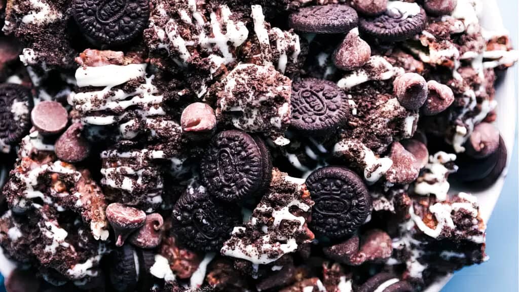 This image shows a close-up of a mix of chocolate-flavored mini cookies, chocolate chips, and drizzled white chocolate. The textures and colors of the ingredients, with dark chocolate and white accents, create a rich and indulgent dessert presentation perfect for snack recipes.