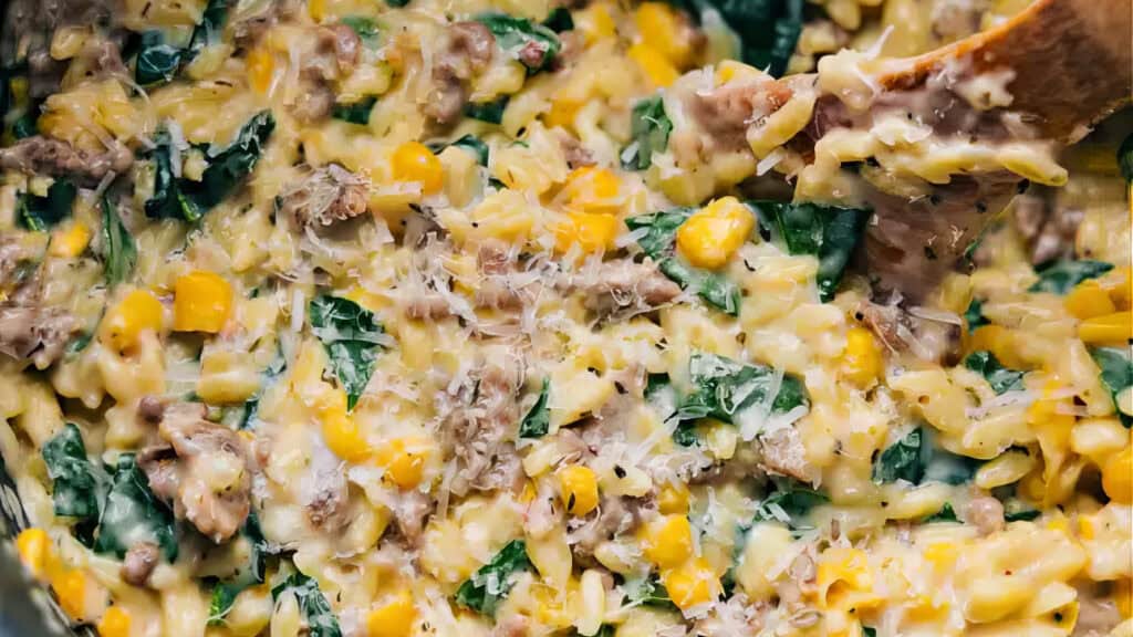 A close-up image of a creamy dish featuring orzo pasta mixed with ground meat, spinach, corn, and grated cheese—a perfect addition to your spinach recipes. All the ingredients are being stirred with a wooden spoon, creating a hearty, cheesy mixture that looks irresistibly delicious.