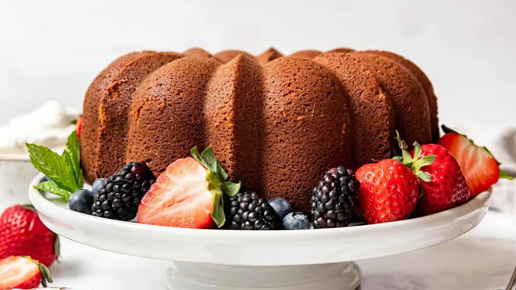 A beautifully baked bundt cake placed on a white cake stand, surrounded by an assortment of fresh berries, including strawberries, blackberries, blueberries, and mint leaves. The cake has a rich brown color and a light dusting of powdered sugar—perfect for showcasing in your favorite Bundt Cake Recipes.