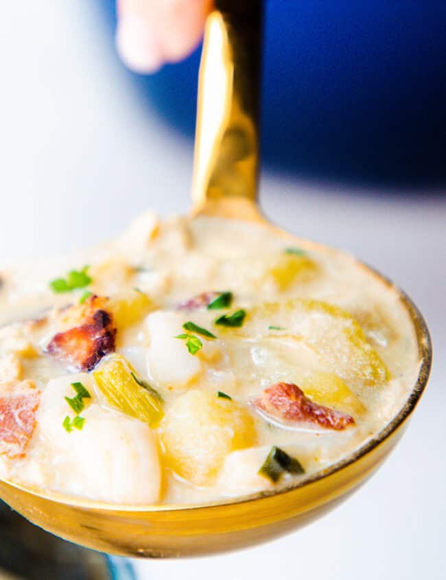 A close-up of a ladle holding a creamy soup with chunks of vegetables and bacon, designed to warm your soul. The dish is garnished with chopped herbs, perhaps parsley. A bowl of this comfort food is partially visible in the background.