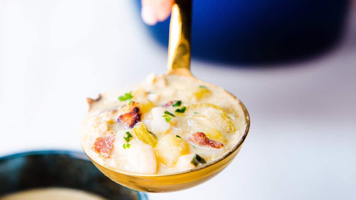 A close-up of a ladle holding a creamy soup with chunks of vegetables and bacon, designed to warm your soul. The dish is garnished with chopped herbs, perhaps parsley. A bowl of this comfort food is partially visible in the background.