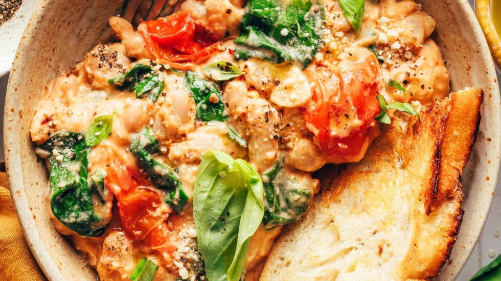 A close-up bowl of creamy tomato gnocchi topped with wilted spinach and fresh basil leaves, perfect for those exploring spinach recipes. The dish is garnished with cracked black pepper and grated cheese. A slice of toasted bread is placed on the side for dipping.