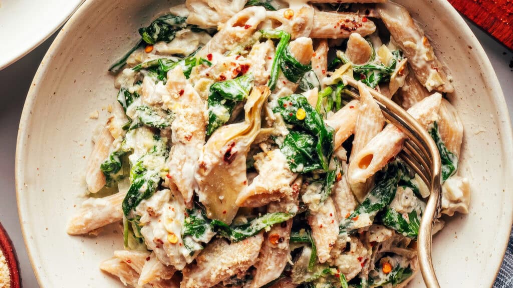 A bowl of creamy pasta with penne and spinach is a delicious addition to your favorite spinach recipes, topped with a light sprinkling of grated cheese. Garnished with red pepper flakes for added flavor, the dish invites you to dig in with a fork placed enticingly on the right side.