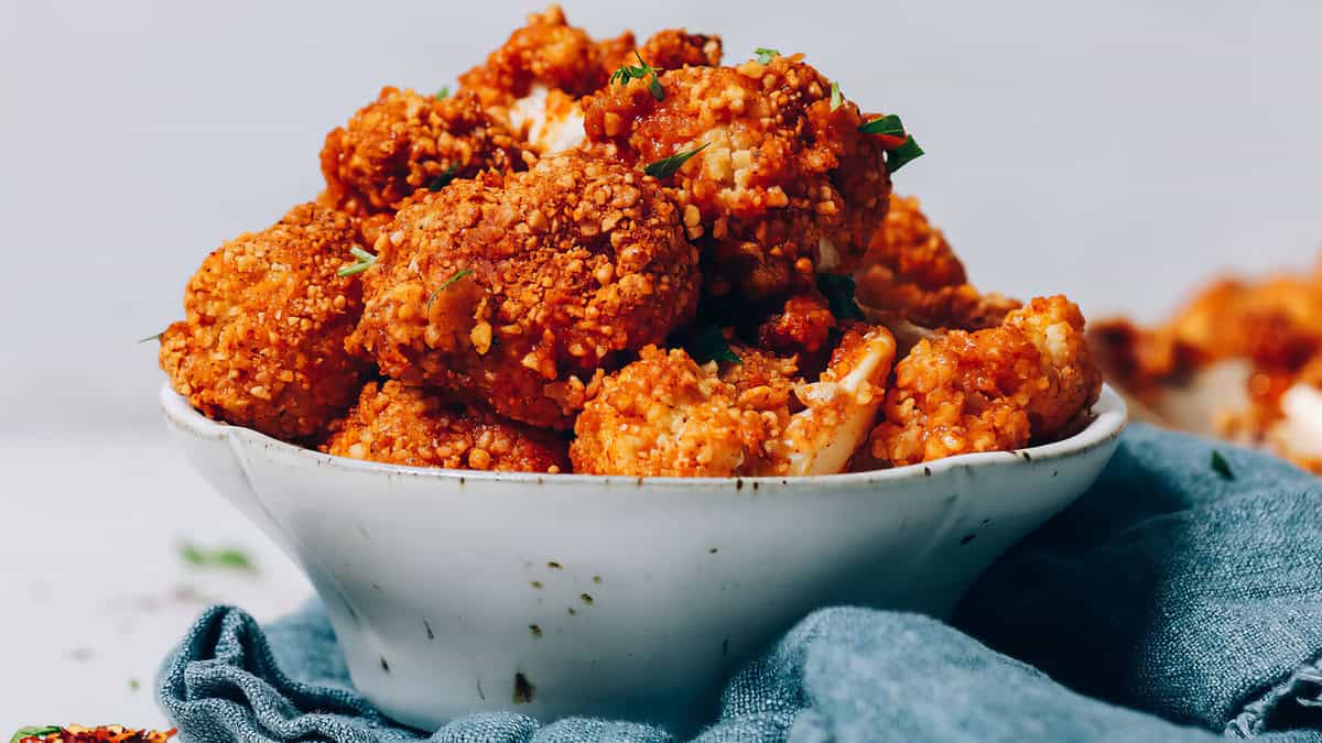 A white bowl filled with golden and crispy cauliflower bites, garnished with chopped herbs, sits on a blue cloth against a light background. The cauliflower, perfect for breaded recipes, appears deliciously seasoned.