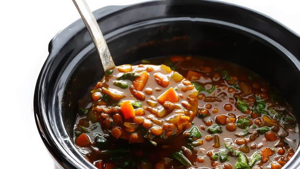 A close-up image of a ladle scooping hearty lentil soup from a black slow cooker. The soup includes visible chunks of carrots, spinach, and lentils in a thick, rich broth—a perfect addition to your collection of slow cooker recipes.