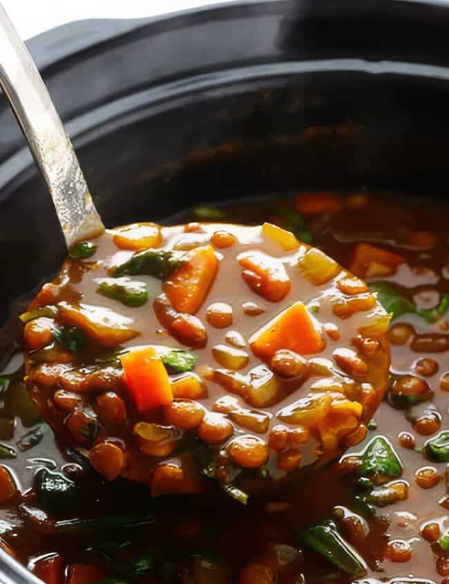 A close-up image of a ladle scooping hearty lentil soup from a black slow cooker. The soup includes visible chunks of carrots, spinach, and lentils in a thick, rich broth—a perfect addition to your collection of slow cooker recipes.