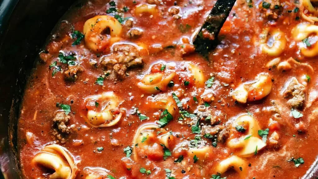 A close-up of a pot filled with a hearty tomato-based soup, featuring cheese tortellini, ground meat, and garnished with fresh parsley. The soup looks thick and flavorful, with a ladle partially submerged in the mixture—perfect for those who love slow cooker recipes.