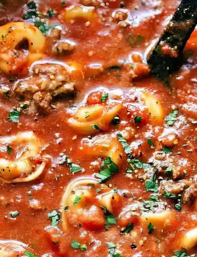 A close-up of a pot filled with a hearty tomato-based soup, featuring cheese tortellini, ground meat, and garnished with fresh parsley. The soup looks thick and flavorful, with a ladle partially submerged in the mixture—perfect for those who love slow cooker recipes.