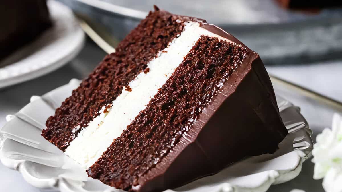 A close-up of a slice of chocolate cake with white frosting between the layers. The cake is covered in a smooth chocolate ganache and is placed on a decorative white plate. This delightful dessert, perfect for your favorite dessert recipes, sits on a slightly blurred gray surface in the background.