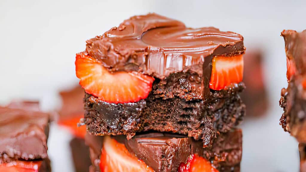 A close-up of a decadent chocolate brownie topped with a glossy layer of chocolate and fresh strawberry slices. The brownie appears moist and rich, with chunks missing from the center, revealing its fudgy interior—a true masterpiece for those seeking the best in Chocolate Recipes. Other brownies are blurred in the background.