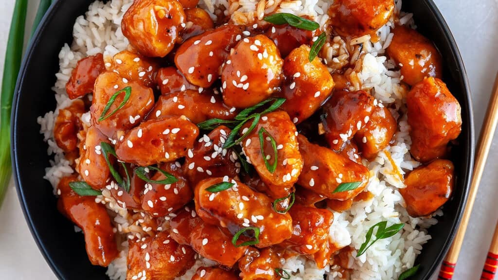 A bowl of white rice topped with glazed chicken pieces garnished with sesame seeds and chopped green onions. The sauce covering the chicken is a rich orange-red color, suggesting a sweet and tangy flavor—a must-try for those exploring new chicken recipes. A pair of chopsticks rests beside the bowl.