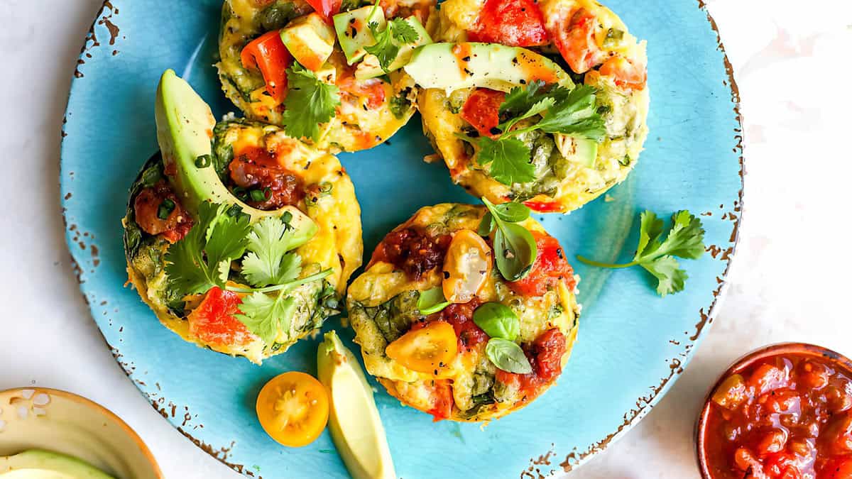 A vibrant breakfast plate with four mini vegetable frittatas garnished with avocado slices, fresh herbs, and tomato salsa. The frittatas are colorful, featuring bell peppers, spinach, and cheese. Surrounding the plate are small dishes containing salsa and cherry tomatoes—an easy egg recipe perfect for lunch or dinner too.