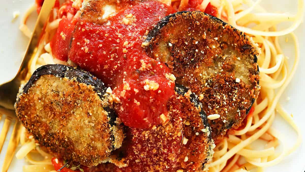 A plate of spaghetti is topped with slices of golden and crispy breaded eggplant and a tomato-based sauce. A fork is placed on the left side of the plate. The dish, one of our delightful recipes, is garnished with grated cheese.