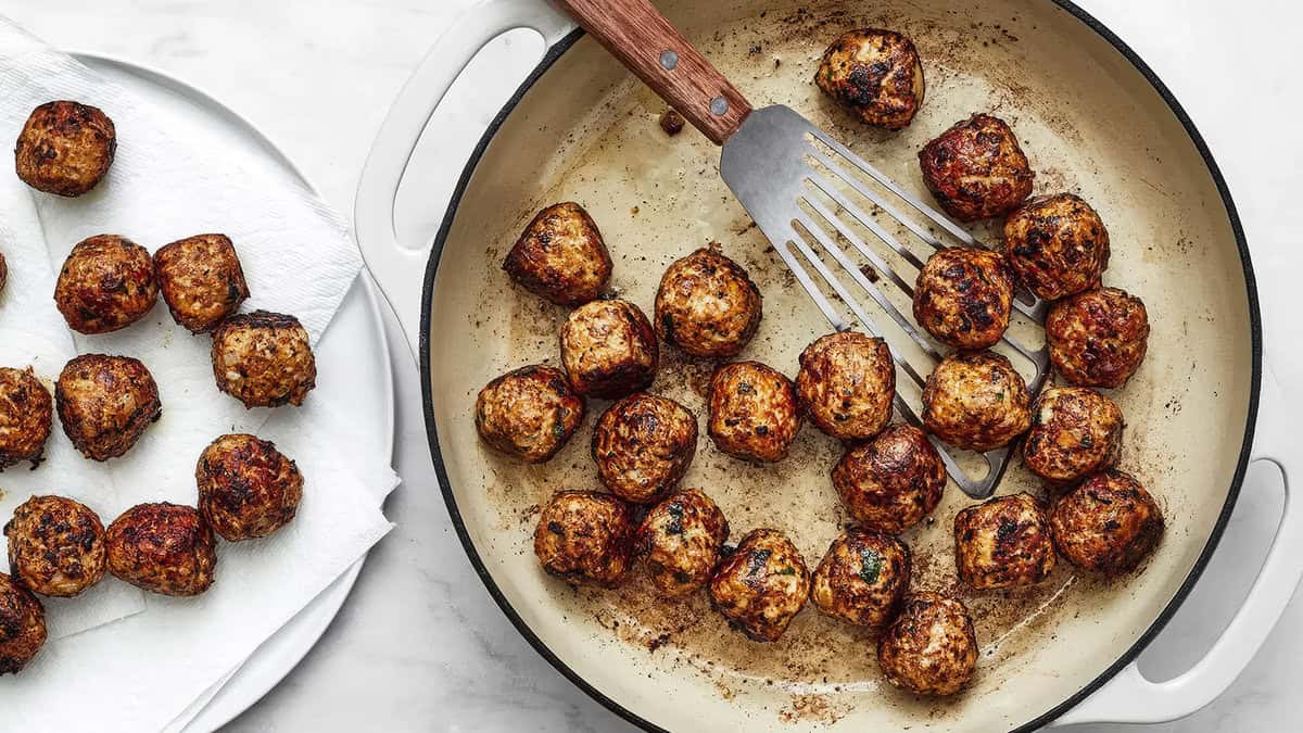 A skillet filled with numerous browned meatballs and a spatula. To the left, a white plate with paper towels has additional cooked meatballs resting on it, showcasing true Meatball Magic. The background surface is a light-colored countertop.