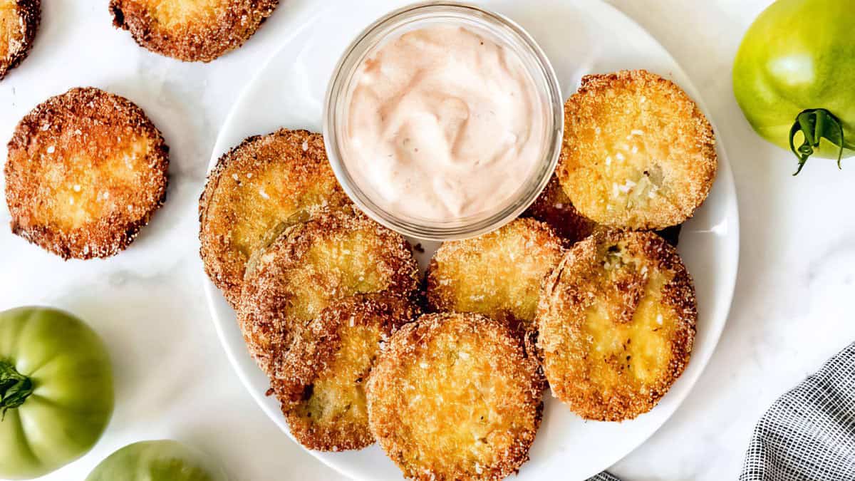 A plate of golden and crispy fried green tomatoes is arranged around a small bowl of creamy dipping sauce. Several green tomatoes are visible nearby, and the background features a white surface and a black and white checkered cloth, making it one of the most delightful breaded recipes you'll try.