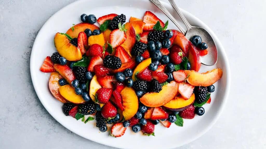 A white oval platter filled with a vibrant mix of fresh fruits, including sliced peaches, strawberries, blackberries, blueberries, and raspberries—an ideal summer dessert. Two spoons rest on the side of the platter, and the background is a light grey surface.