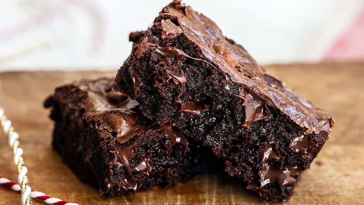 Two gooey brownies stacked on a wooden surface. The rich, dark cocoa color and melty chocolate chunks make them perfect for chocoholics. With a shiny, slightly cracked top, their texture and decadence are highlighted against a softly blurred background.