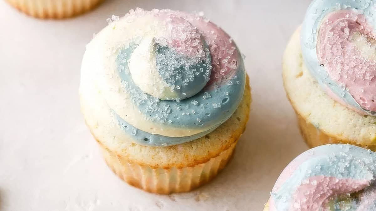 A close-up image of a frosted cupcake features icing swirled in pastel colors of blue, pink, and white, sprinkled with coarse sugar. The cupcake is set on a light-colored surface, with other similarly decorated cakes partially visible in the background. Perfect for any frosting enthusiast's recipes!