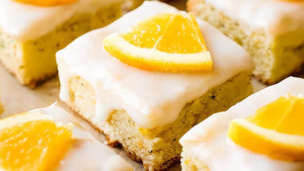 Close-up of several square lemon cake pieces topped with white glaze and garnished with a small orange slice. The moist cakes and glossy icing create a tempting and fresh appearance, perfect for a sweet treat inspired by classic sheet cake recipes.