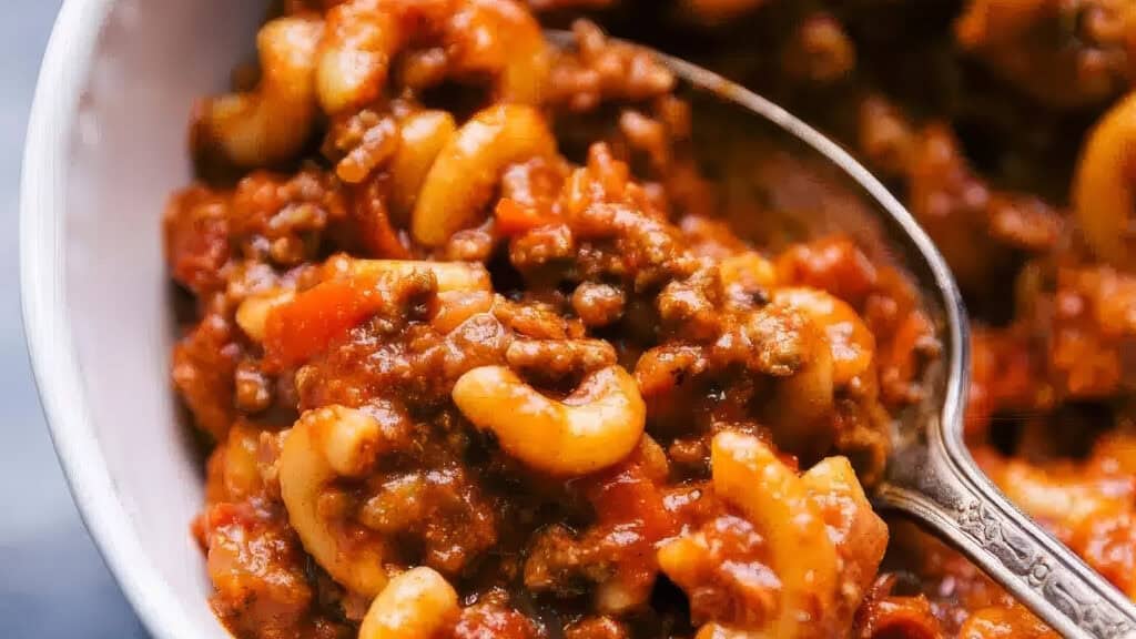 A bowl filled with hearty American goulash, featuring elbow macaroni, ground beef, and a rich tomato-based sauce. A spoon is partially submerged in the dish, ready for serving. This comforting mixture showcases a blend of pasta and savory meat sauce perfect for ground beef recipes enthusiasts.