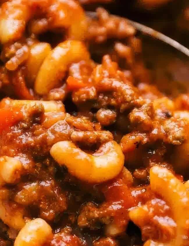 A bowl filled with hearty American goulash, featuring elbow macaroni, ground beef, and a rich tomato-based sauce. A spoon is partially submerged in the dish, ready for serving. This comforting mixture showcases a blend of pasta and savory meat sauce perfect for ground beef recipes enthusiasts.