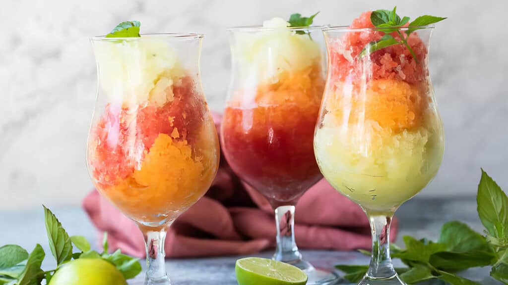 Three colorful sorbet glasses sit on a table, each filled with layered scoops of red, orange, and yellow sorbet. Garnished with fresh mint leaves, the glasses are surrounded by lime halves and mint sprigs, evoking a tableau worthy of sophisticated cocktail recipes. A red cloth rests in the background.