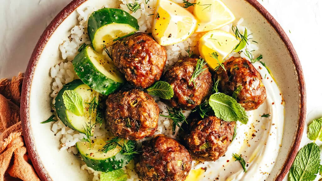 A bowl of rice topped with meatballs, cucumber slices, lemon wedges, and fresh herbs such as mint and dill—a true highlight among ground beef recipes. A creamy white sauce is spread on the side. An orange cloth napkin is off to the side of the bowl.