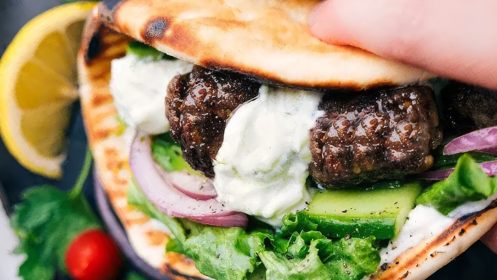 Close-up of a grilled flatbread sandwich filled with seasoned meat, fresh lettuce, cucumber slices, red onions, and topped with a dollop of creamy white sauce. A lemon wedge, cherry tomato, and cilantro garnish are visible in the background—truly a Meatball Magic creation.