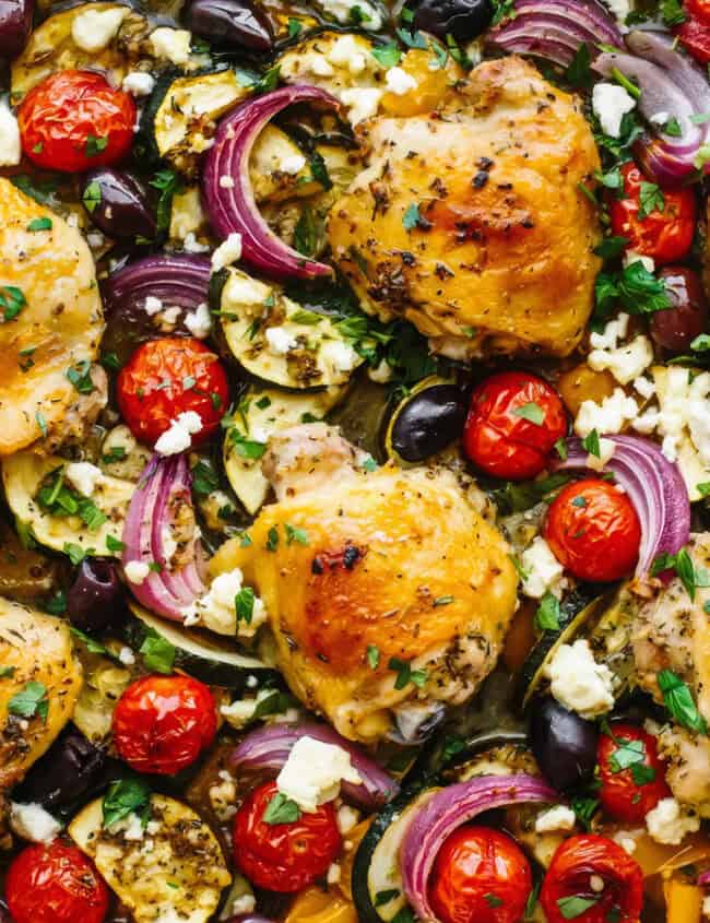 A colorful, appetizing sheet pan meal featuring roasted chicken thighs, cherry tomatoes, red onion slices, zucchini, and bell peppers. Among the finest chicken recipes, the dish is garnished with herbs, olives, and crumbled feta cheese, creating a vibrant and textured presentation.