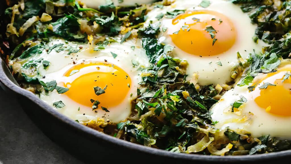 Close-up of a skillet containing baked eggs with runny yolks nestled in a bed of sautéed leafy greens. The greens are garnished with fresh herbs and seasonings, reminiscent of vibrant pancake recipes. The dish showcases bright yellow egg yolks contrasting with the vibrant green vegetables.