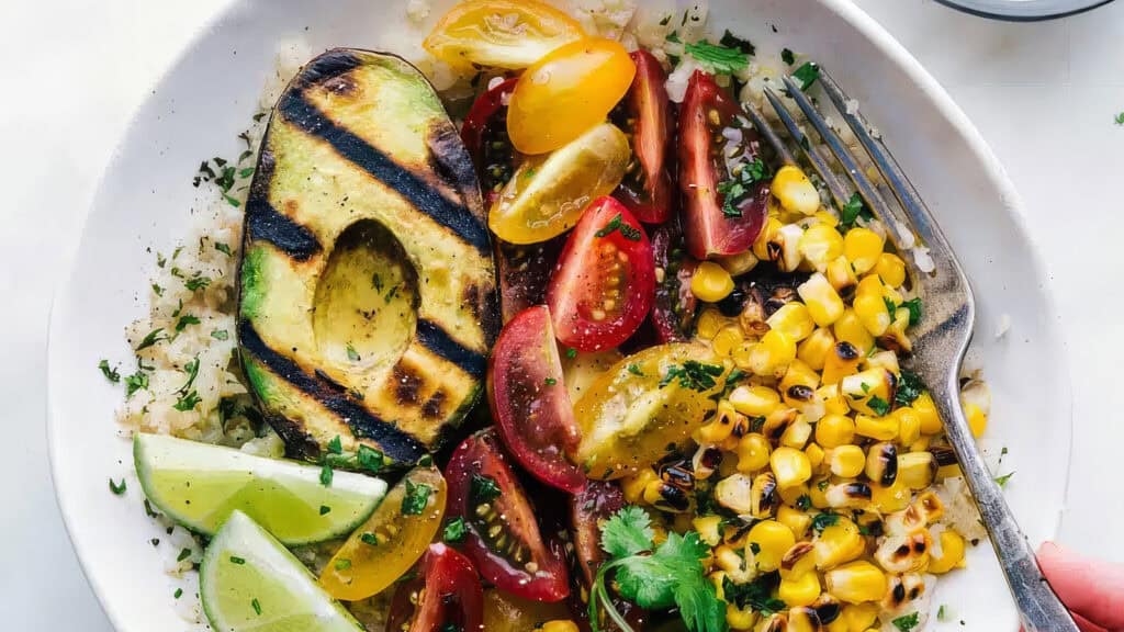 A vibrant bowl filled with grilled avocado, cherry tomatoes, corn kernels, and lime wedges on a bed of rice, garnished with herbs. This healthy dish showcases the fruit's creamy texture. A fork rests on the side of the dish, ready to help enjoy the colorful meal.