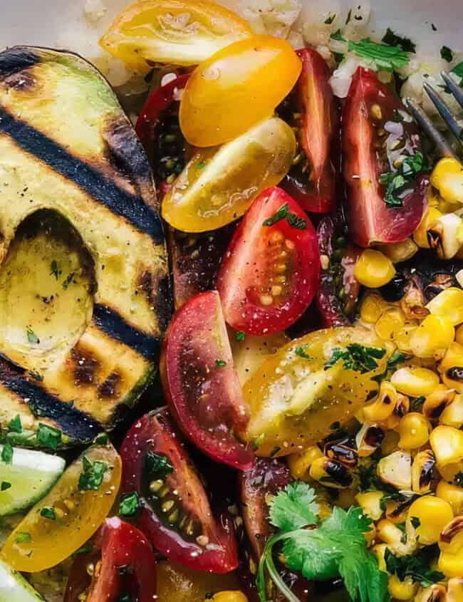 A vibrant bowl filled with grilled avocado, cherry tomatoes, corn kernels, and lime wedges on a bed of rice, garnished with herbs. This healthy dish showcases the fruit's creamy texture. A fork rests on the side of the dish, ready to help enjoy the colorful meal.