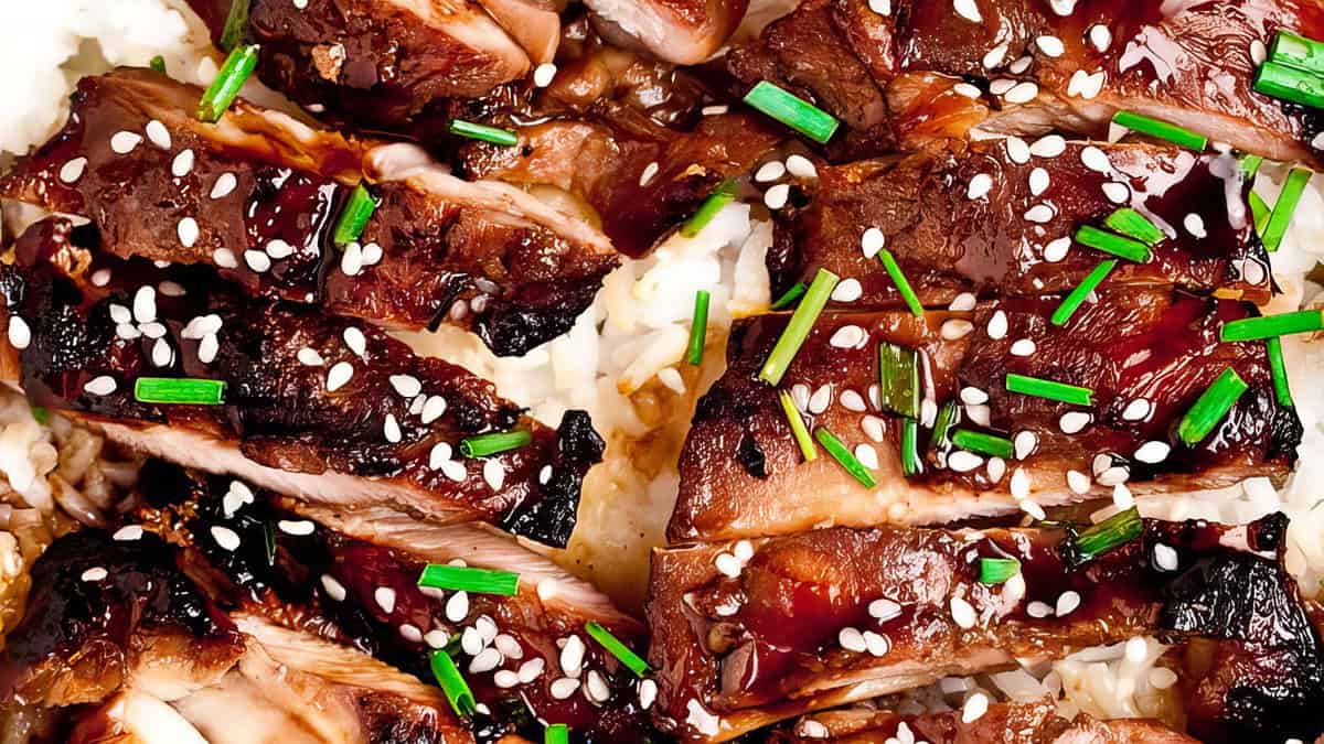 Close-up of succulent beef teriyaki topped with sesame seeds and garnished with chopped green onions, served over a bed of white rice. This easy recipe is perfect for back-to-school lunches; the beef is glazed with a rich, dark sauce, giving it a glossy appearance. The dish looks savory and appetizing.