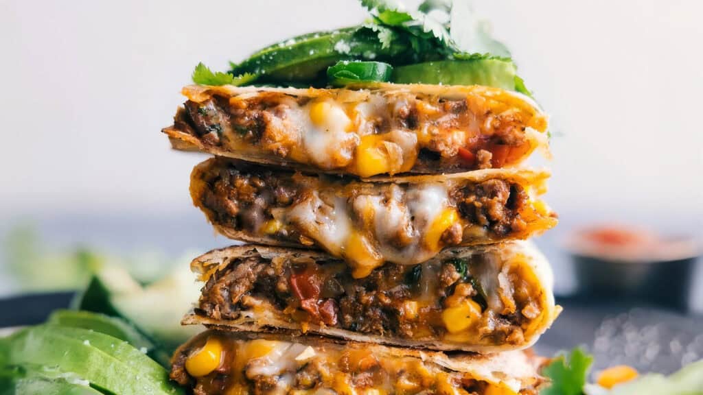 A close-up shot of a stack of cheesy, beef and vegetable quesadillas. The layers reveal ground beef mixed with corn, tomatoes, and melted cheese. Fresh cilantro and sliced jalapeños garnish the top, while slices of avocado sit to the side—perfect for ground beef recipes enthusiasts.