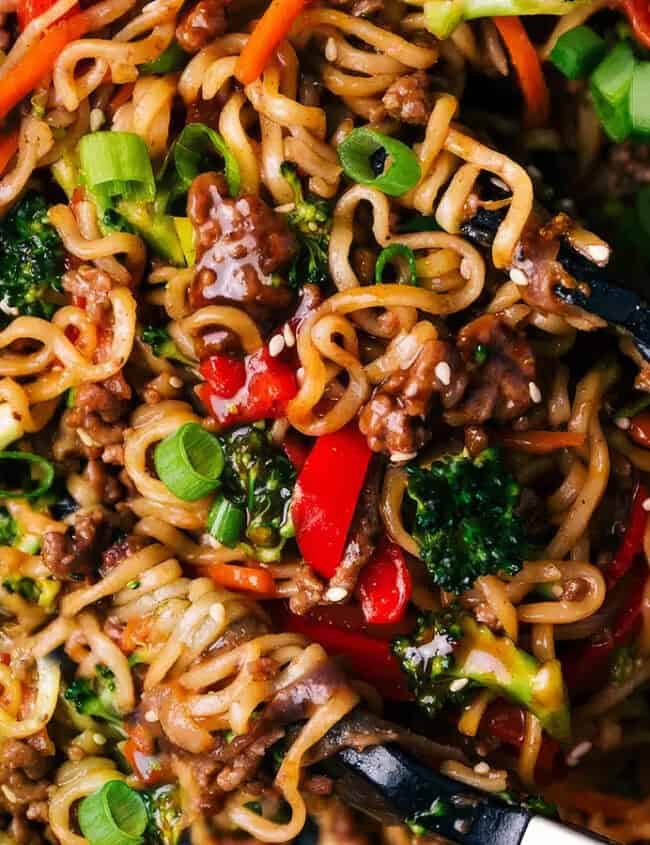 A close-up shot of a vibrant noodle stir-fry dish filled with colorful vegetables like red bell peppers, broccoli, and spring onions, mixed with chunks of ground beef and garnished with sesame seeds. The food is being served with a pair of tongs, showcasing one of the tastiest ground beef recipes.