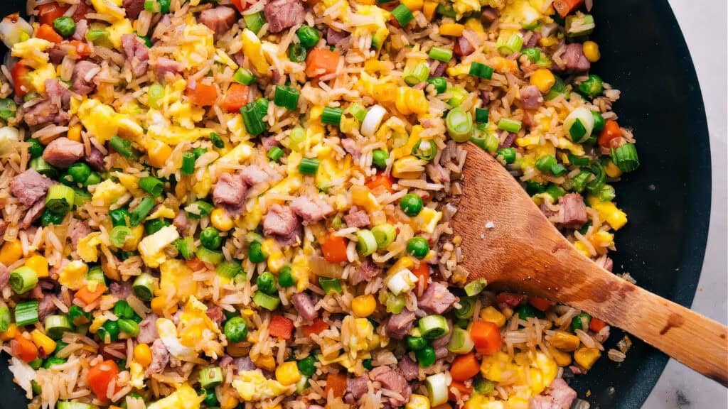 A close-up view of a colorful fried rice dish in a pan, featuring diced beef, corn, peas, carrots, and chopped spring onions. A wooden spoon is partially visible, resting on the right side of the pan. The ingredients are well-mixed and look freshly cooked—perfect for any rice dish recipes collection.