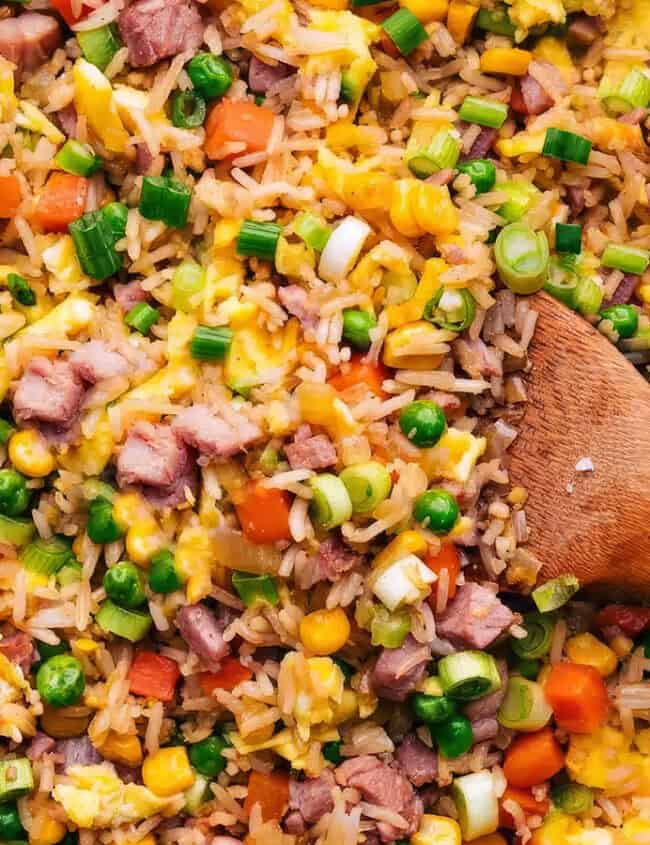 A close-up view of a colorful fried rice dish in a pan, featuring diced beef, corn, peas, carrots, and chopped spring onions. A wooden spoon is partially visible, resting on the right side of the pan. The ingredients are well-mixed and look freshly cooked—perfect for any rice dish recipes collection.