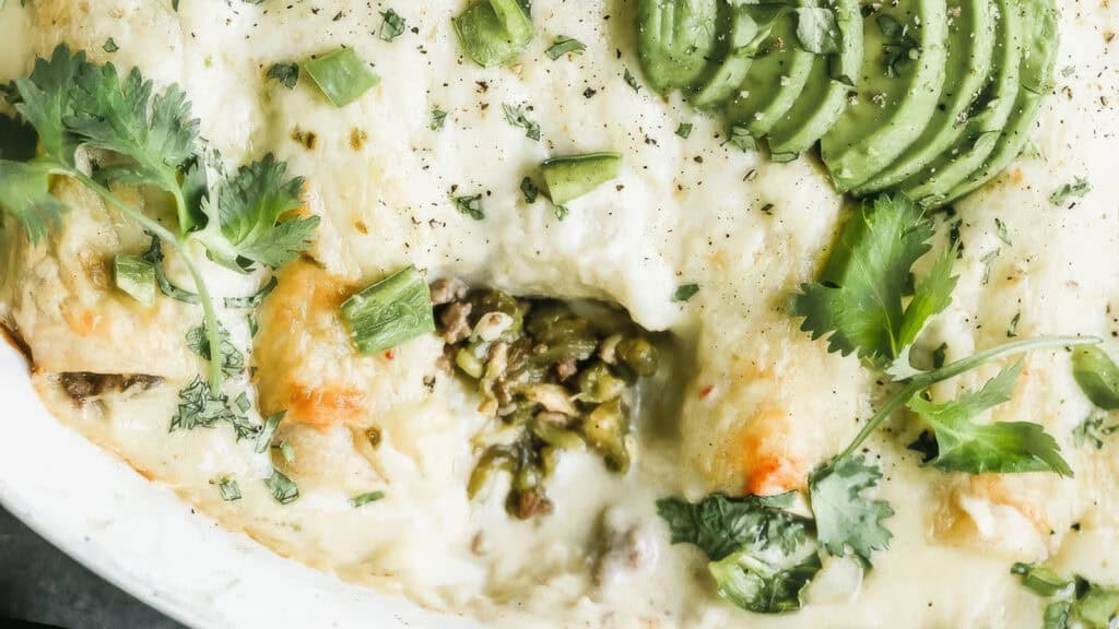 A close-up of a baked dish covered with melted cheese and garnished with slices of avocado, green onions, and cilantro. A portion has been taken out, revealing a filling of minced meat and green chilies—a perfect addition to your favorite ground beef recipes.